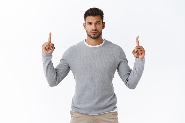 Skeptical unsure handsome macho man in grey sweater pointing up and looking hesitant camera raise one eyebrow arched as feeling kinda suspicious and hesitant white background