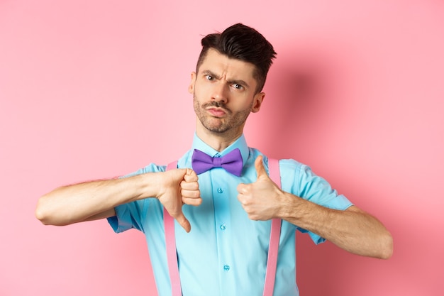 Skeptical young man in bow-tie grimacing, showing thumbs up and down, standing indecisive and judging something, standing over pink background.