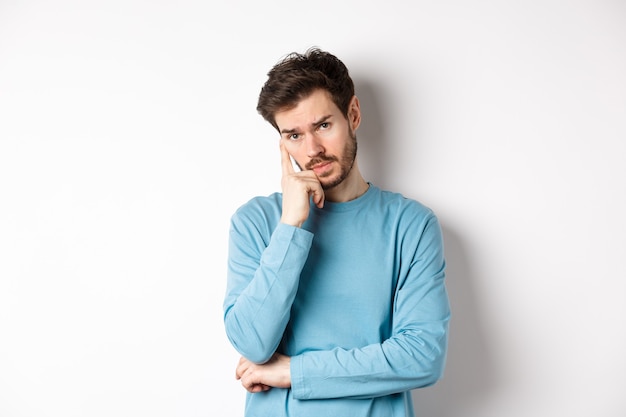 Skeptical young man listening to you with bored face, looking at camera reluctant, standing in sweatshirt over white background