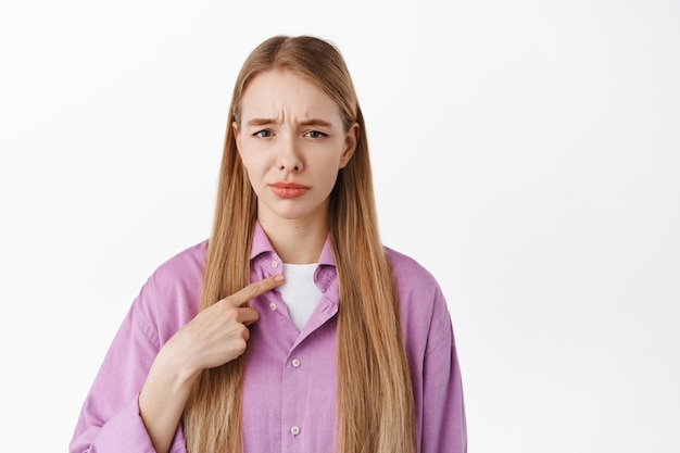 Skeptical young woman grimacing, pointing at herself with unconfident doubtful expression, standing against white wall