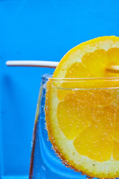 Skewered orange dangling in fizzy glass of bubbly soda on blue background