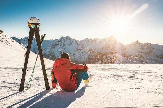 晴れた日に雪山に座っているスキーヤー選手-彼の隣に空のギアで夕日を楽しむ大人の男-冬のスポーツと休暇の概念-男性の体に焦点を当てる