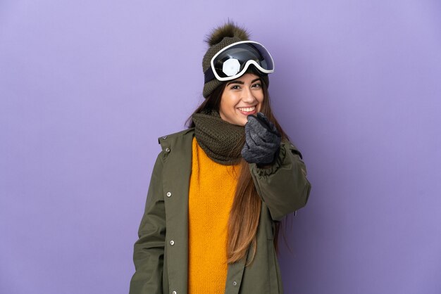 Skier caucasian girl with snowboarding glasses isolated on purple background making money gesture