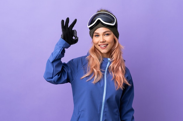 Skier girl with snowboarding glasses on isolated purple showing ok sign with fingers