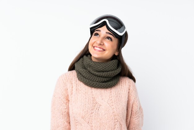 Skier girl with snowboarding glasses over isolated white wall looking up while smiling