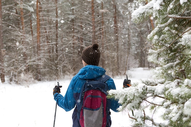 Skiër met een rugzak en muts met pompon met skistokken in zijn handen op de achtergrond van een besneeuwd bos. Langlaufen in het winterbos, buitensporten, gezonde levensstijl, wintersporttoerisme.