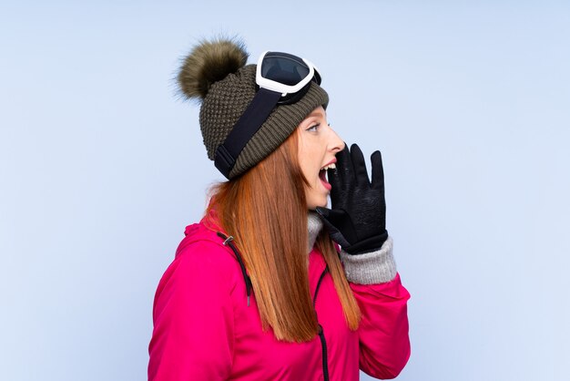 Фото Рыжая женщина лыжник в сноуборд очки над синей стеной, крича с широко открытым ртом