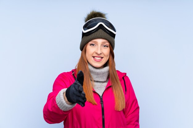 Skiër roodharigevrouw met snowboarding glazen over geïsoleerd blauw muurhandenschudden na goede deal