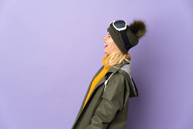 Skier Russian girl with snowboarding glasses isolated on purple background laughing in lateral position