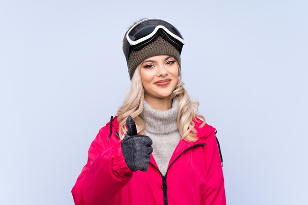 Девушка подростка лыжника с стеклами сноубординга над изолированной синью давая жест больших пальцев руки вверх