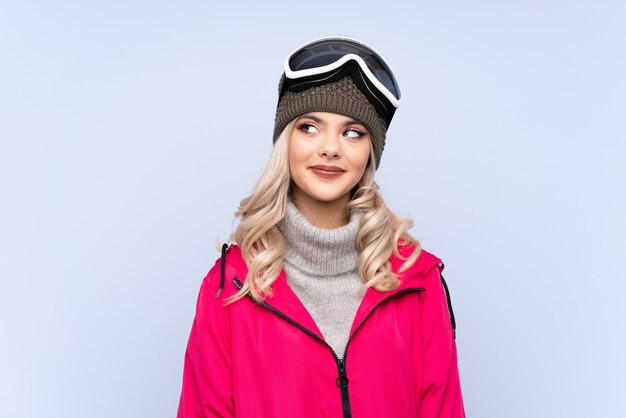 Photo skier teenager girl with snowboarding glasses over isolated blue  laughing and looking up