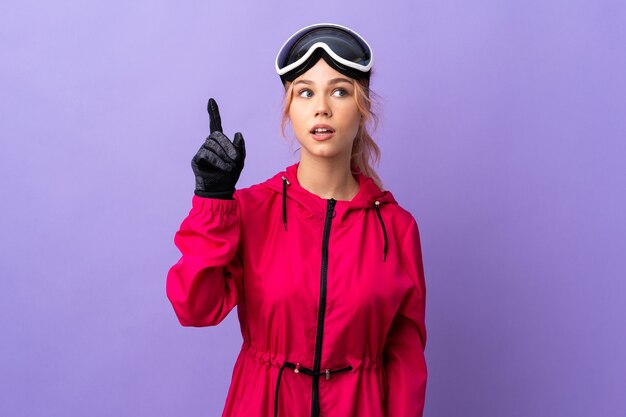 Skier teenager girl with snowboarding glasses over isolated purple  intending to realizes the solution while lifting a finger up