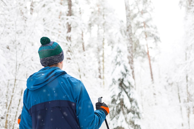 Skier in windbreaker and hat with pompom with ski poles in his hands with his back on a snowy forest. Cross-country skiing in winter forest, outdoor sports, healthy lifestyle.