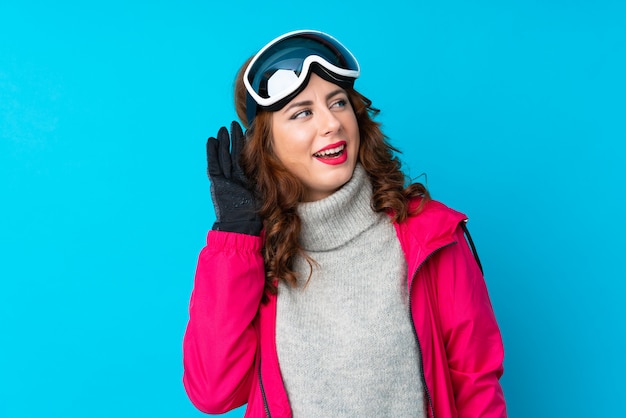 Skier woman with snowboarding glasses over isolated blue wall listening something