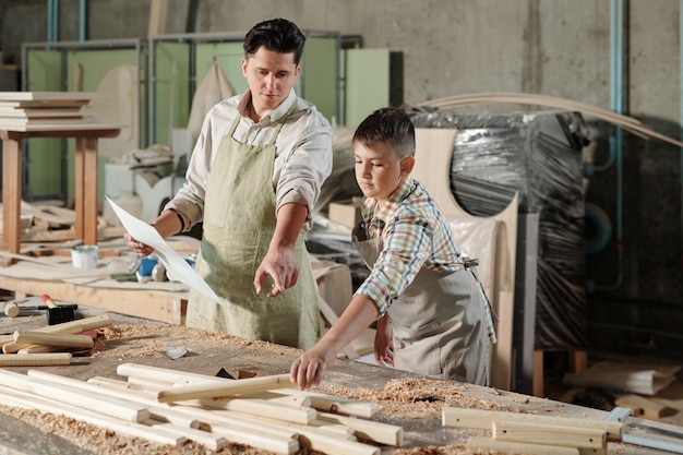 Skilled carpenter in apron pointing at wooden detail while teaching son to make furniture in workshop