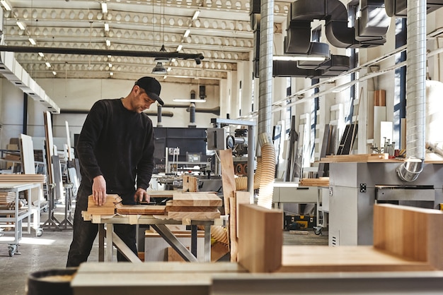 Abile falegname in uniforme nera che taglia un pezzo di legno nel suo laboratorio di falegnameria