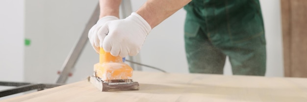 Skilled carpenter polishes wooden surface of office table with grinder machine man in