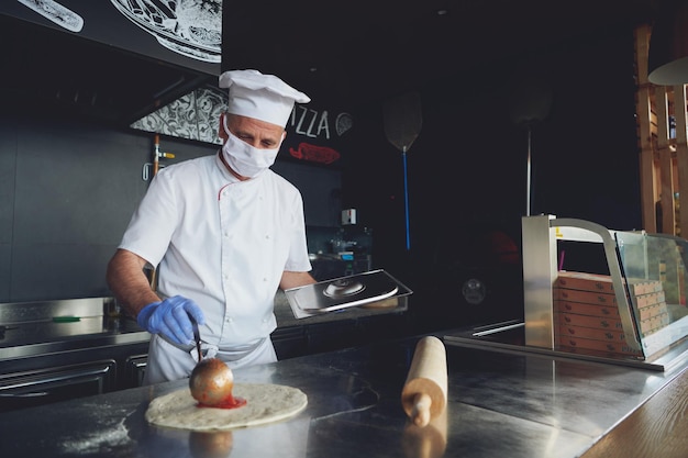 특별한 장작불 오븐이 있는 현대적인 레스토랑 주방 내부에서 숙련된 요리사가 전통 이탈리아 피자를 준비하고 있습니다. 코로나바이러스 new normal conce에서 보호용 의료용 안면 마스크와 장갑 착용