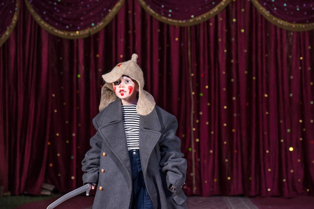 Photo skilled empathetic boy pretending to be a brave russian soldier while performing a dramatic act, on stage, against dark red classical curtains