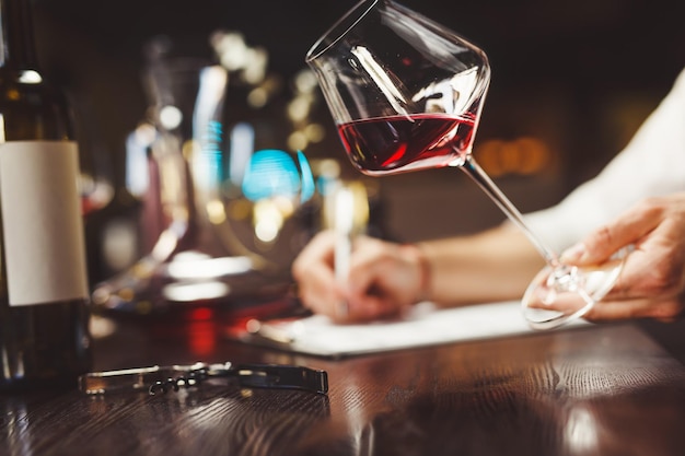 Skilled expert writes on paper holding elegant wineglass of expensive red wine at table in luxury restaurant close view