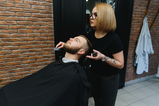 Skilled female barber cutting a beard of brunette adult guy in professional retro barbershop.