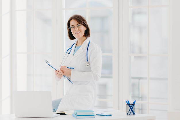 Skilled female therapist or cardiologist writes information at clipboard ready to consult and cure patients wears white coat poses at hospital office with laptop computer uses phonendoscope