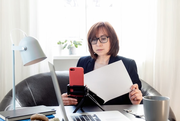 Skilled middle aged woman communicating online or distance studying by phone on the tripod,