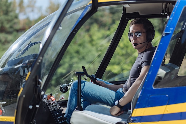 Skilled professional. Athletic young pilot sitting at a pilot booth of a helicopter and posing for the camera, being ready to start a flight