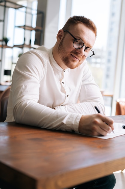 Skilled young songwriter wearing stylish eyeglasses is writing note of song with pen