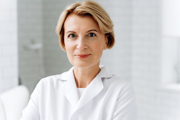 Skillful specialist. Portrait of mature cosmetologist in white lab coat looking at camera with smile while posing at her clinic