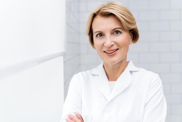 Skillful specialist Portrait of mature cosmetologist in white lab coat looking at camera with smile while posing at her clinic