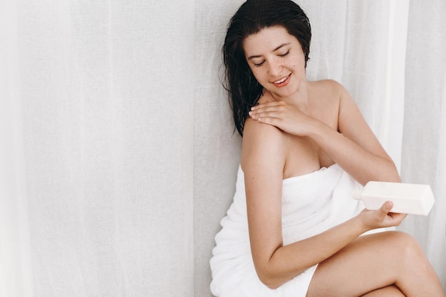 Photo skin and body care happy sexy woman in white towel applying moisturizing cream on shoulder in bathroom slim young woman relaxing spa and wellness hand holding plastic bottle with lotion