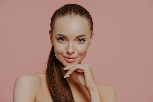 Уход за кожей и антивозрастные процедуры Уверенная брюнетка держит руку под подбородком, расчесала волосы, чистая свежая кожа наслаждается косметическими процедурами, позирует с обнаженным телом на розовом фоне