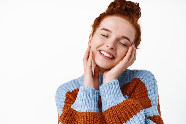 Skin care beauty. Smiling redhead woman touching her glowing natural facial skin without blemished, no make up healthy skin, close eyes and stand delighted on white