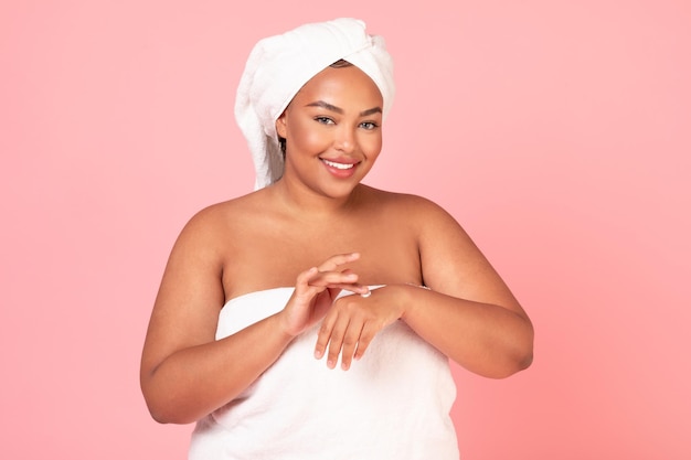 Skin care cosmetics concept Excited oversized black lady applying moisturizer cream on hands standing wrapped in towel