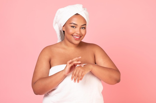 Фото Концепция косметики по уходу за кожей взволнованная негабаритная чернокожая дама наносит увлажняющий крем на руки, завернутые в полотенце