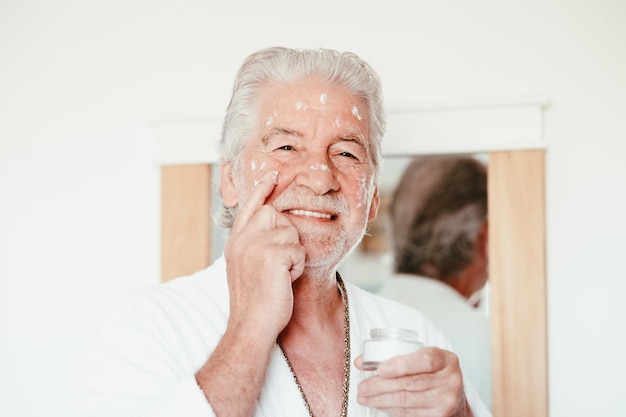 Skin care Handsome elderly bearded man in bathrobe applying cream on face as daily routine holding a jar of cream