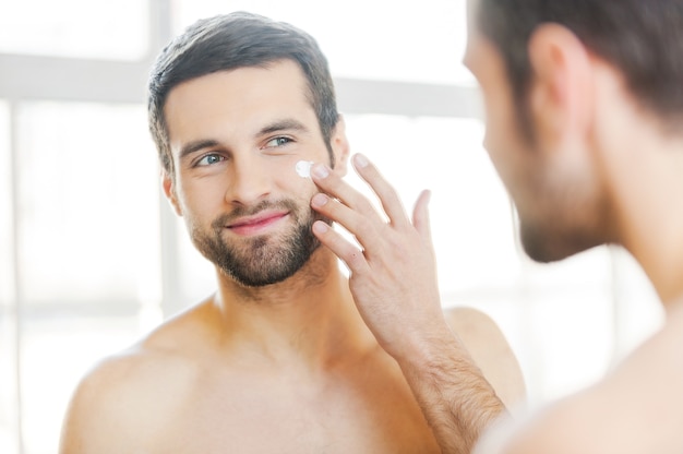 Photo skin care. handsome young shirtless man applying cream at his face and looking at himself with smile while standing in front of the mirror