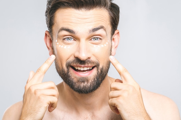 Skin care. Handsome young shirtless man applying cream at his face and looking at himself with smile while standing over gray background