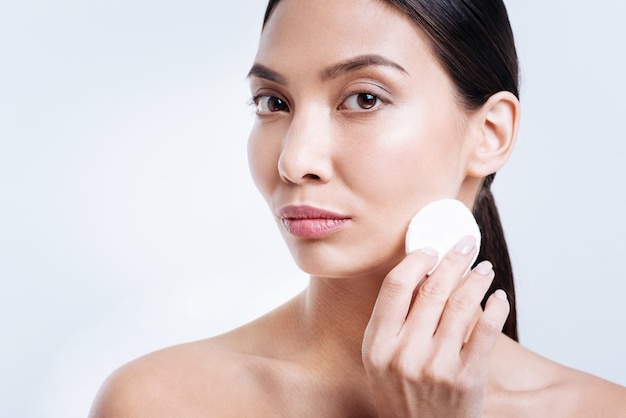 Skin care. Pleasant dark-haired young woman with bare shoulders holding a cotton pad near her face, being about to remove makeup, while standing isolated on a white wall