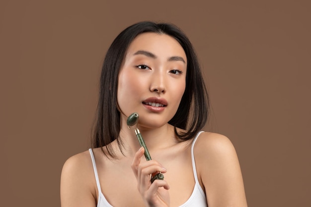 Skin care treatments korean lady using greenstone jade facial roller posing over brown studio