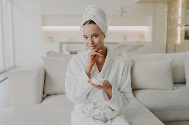 Концепция ухода за кожей и красоты. портрет молодой привлекательной женщины в халате и полотенце на голове, держащей банку с кремом и улыбающейся, сидя на диване в гостиной, делая косметические процедуры