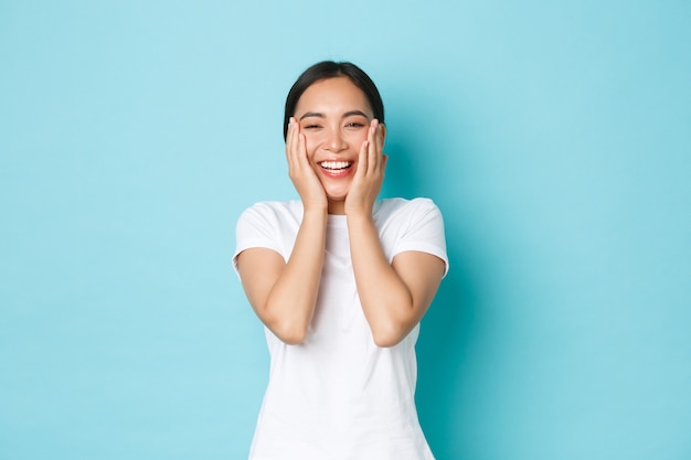 Concetto di cura della pelle, bellezza e stile di vita. ragazza asiatica sorridente allegra che si rallegra, sembra felice, tocca la pelle perfettamente pulita e si rallegra, si è sbarazzata dell'acne, in piedi muro blu