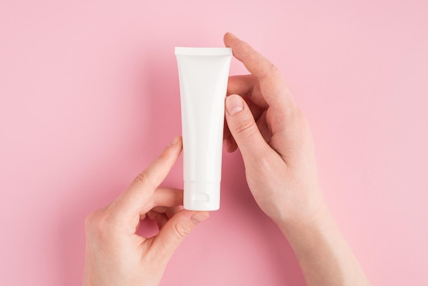 Skincare concept. Top above overhead pov first person view photo of female hands holding white tube of cream isolated on pastel pink background with copyspace