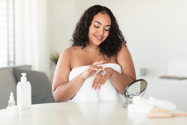 Photo skincare cosmetics concept african american plus size lady applying hands cream sitting at dressing table in bedroom