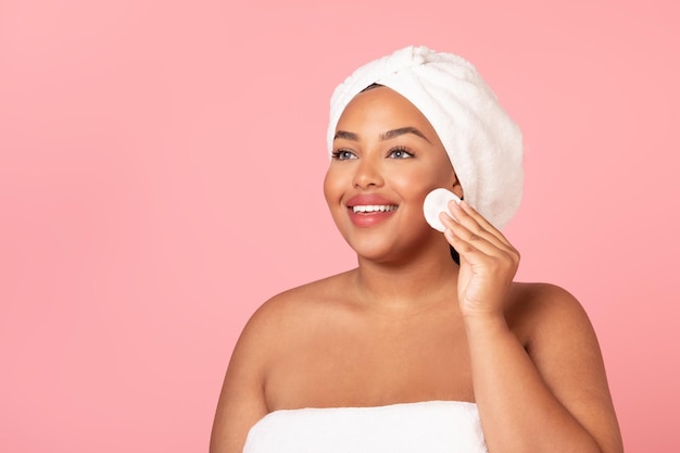 Skincare routine Happy black chubby woman cleansing skin with cotton pad removing makeup from face pink background