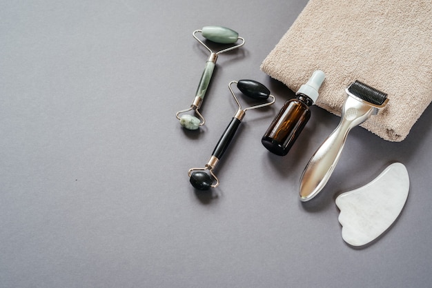 Инструменты по уходу за кожей: дерма-валик с микроиглами, массажные валики из нефрита-гуаши и флакон с сывороткой на сером фоне.