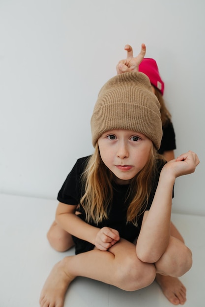 Skinny boy in a beige knitted hat lookig at the camera. His siter is giving him bunny ears, hiding behind him