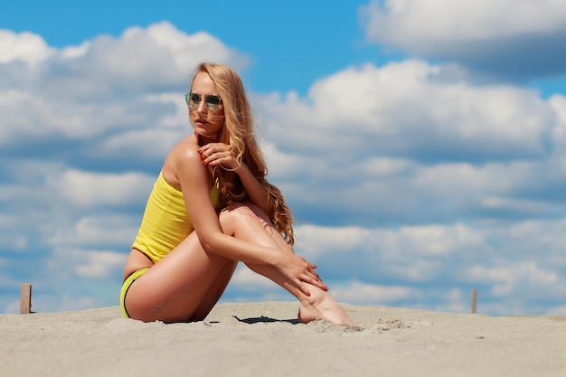 Skinny long haired sexy woman with sun glasses posing in seductive energetic posture in sandy beach on background of cloudy sky