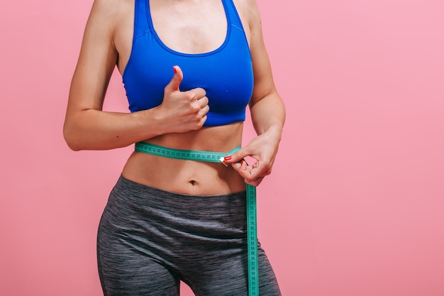 Skinny woman measures waist and shows thumb on pink wall